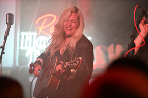 A blonde woman performing country music on stage while playing an acoustic guitar, with a microphone and a blurred background featuring a neon sign for Ron's Honky Tonk.