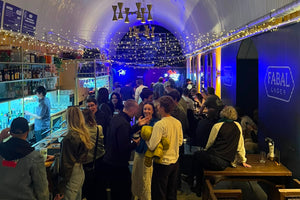 A crowded indoor venue with a curved white ribbed ceiling, hanging gold chandeliers, blue ambient lighting, and a bar counter on the left. Several people are standing and sitting at wooden tables on the right, and a visible sign reads FABAL LONDON.