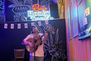 A man stands on a small wooden stage playing an acoustic guitar in front of a black backdrop with white floral drawings and a glowing neon sign that reads 'Ron's Honky Tonk.' The scene is lit with purple and warm orange lighting, and a speaker, chair, and stage equipment are visible.