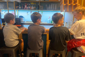 Four boys sitting on stools at a bar, viewed from behind as they watch the bartender prepare drinks. The setting features wooden shelving with glassware and bottles in the background at ron's honky tonk